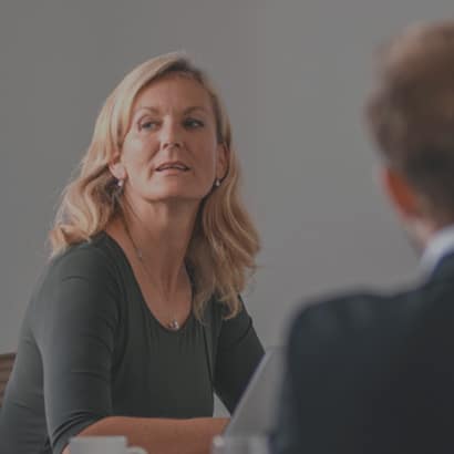 A older business woman looking off the left. She is white, has blonde hair and is wearing a green top.