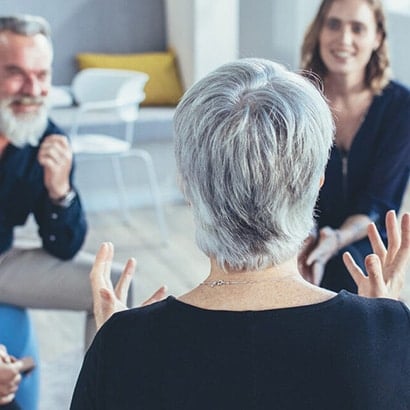 An older woman with short grey hair, wearing a dark blue jumper, facing away from the camera and having a conversation with two other people. One is an older white man with grey hair and a beard, the other is a young white woman with blonde curly hair and a blue zip up top.