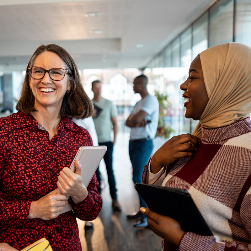 Two women holding iPads, smiling. One is a black women in a hijab, wearing a jumper. The other is a white woman, wearing glasses and a patterned shirt.