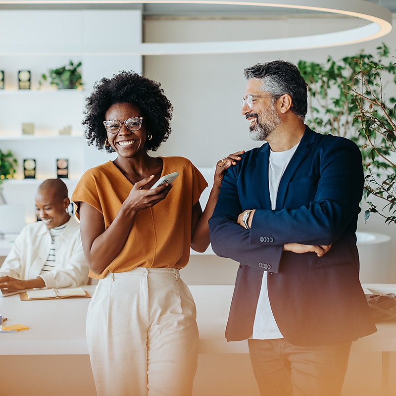 Two colleagues having a conversation and laughing. One is a middle aged black woman, wearing glasses and an orange top, holding a phone and smiling. The other is a middle aged man with a beard and glasses, wearing a white t-shirt under a blue blazer.