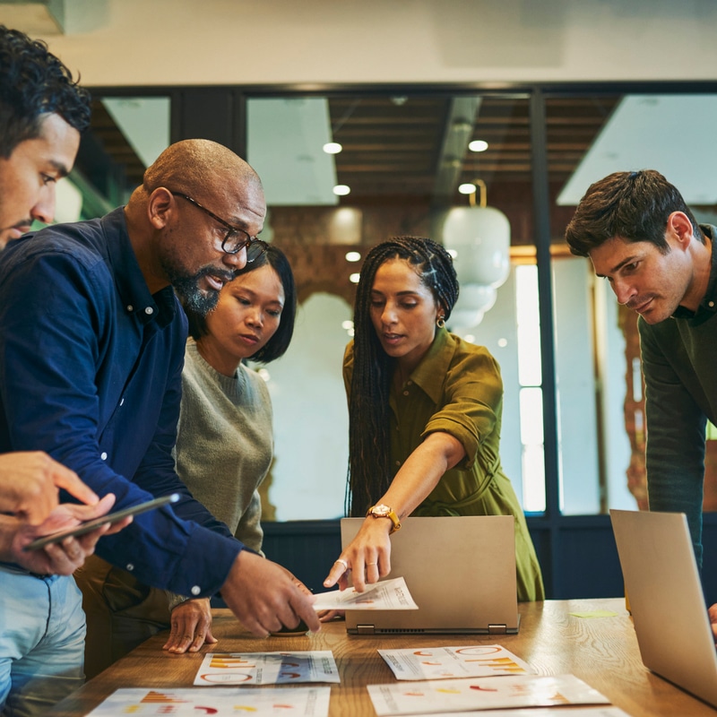 Colleagues meeting around a desk, standing up and looking at documents on the table. There is a group of five colleagues of mixed ethnicities, three men and two women.