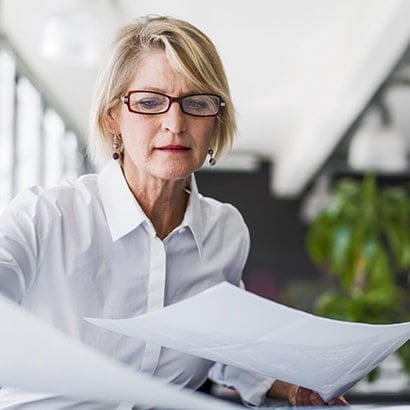 An older white business woman holding a document and looking at another document. She is blonde, wearing glasses and a white shirt.