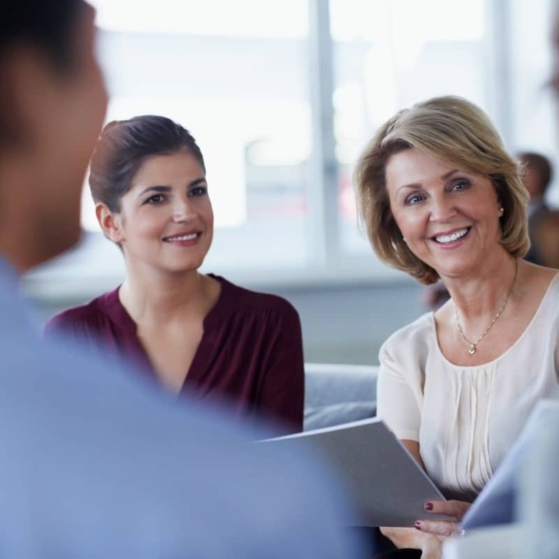 Two business women in a conversation with others. One is blonde and slightly older, the other has scraped back dark hair.