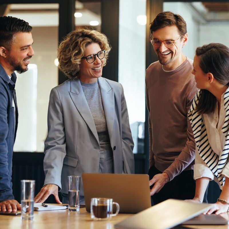 A man in a blue shirt, a women in a light blue suit, a man in a light maroon top and a women in a white shirt, all standing around a laptop