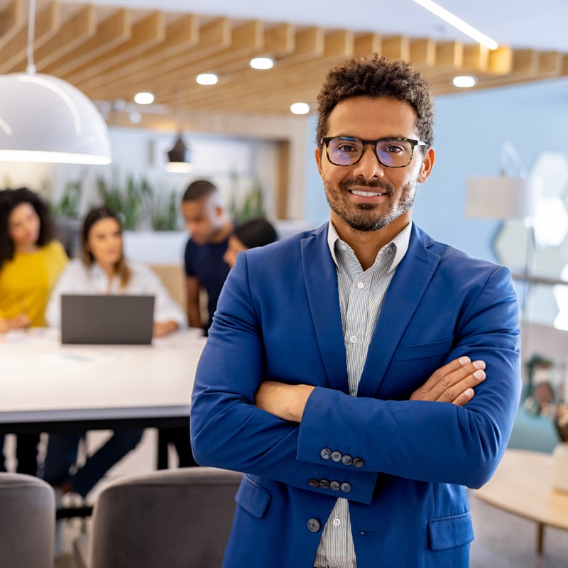 Young, black, male professional smiling to the camera, wearing glasses and a blue blazer with an open collar shirt on. His colleagues are working behind him.